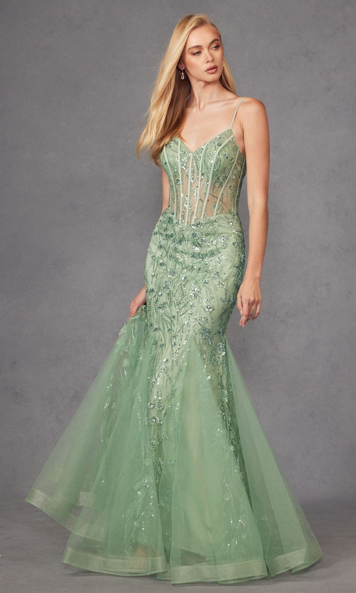 A woman models the Juliet JT2441S long sequin mermaid prom dress with spaghetti straps in light green, standing against a gray backdrop and elegantly holding the skirt.